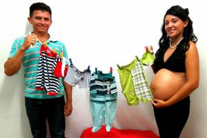 Foto de uma mulher grávida e o pai segurando um varal com roupinhas de recem-nascido