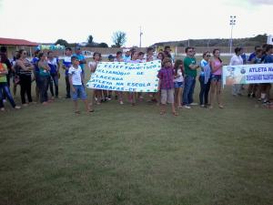 Alunos da Escola Francisco Elianúbio de Lacerda com cartaz de identificação da escola no campo de futebol participando do programa Atleta na Escola